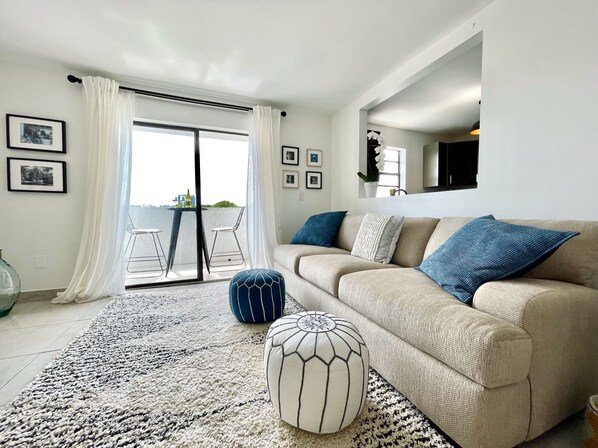 A modern living room with a beige sofa, blue and white cushions, a patterned rug, and a glass door leading to an outdoor balcony.
