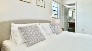 A neatly made bed with white linens and patterned pillows, a framed picture on the wall, and a bathroom visible through an open door.