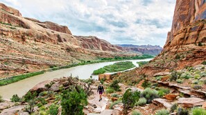 A hiker on a rocky trail with a river and mountains in the background. - Entrada at Moab 594 (Moab)