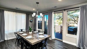 Un comedor con una mesa de madera dispuesta para seis personas, rodeada de sillas. Encima de la mesa, un candelabro, y la habitación tiene ventanas amplias con cortinas.