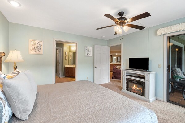 A bedroom with a bed, a ceiling fan, a fireplace, a television, and a door leading to a bathroom. - Wimbledon Beauty | Cozy and Charming (Branson)