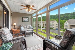 A covered porch with wicker furniture, a ceiling fan, and large windows offering a view of residential buildings. - Wimbledon Beauty | Cozy and Charming (Branson)