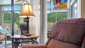 A cozy room with a wicker chair, a table with a lamp, and a view of greenery through large windows. - Wimbledon Beauty | Cozy and Charming (Branson)
