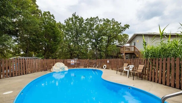 A swimming pool with a wooden fence, lounge chairs, and a building in the background. - Enchanted Woods Retreat / Pools / Hot Tub (Branson)