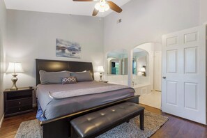 A bedroom with a bed, nightstands, a bench, a ceiling fan, a painting, and a bathroom visible through an archway.