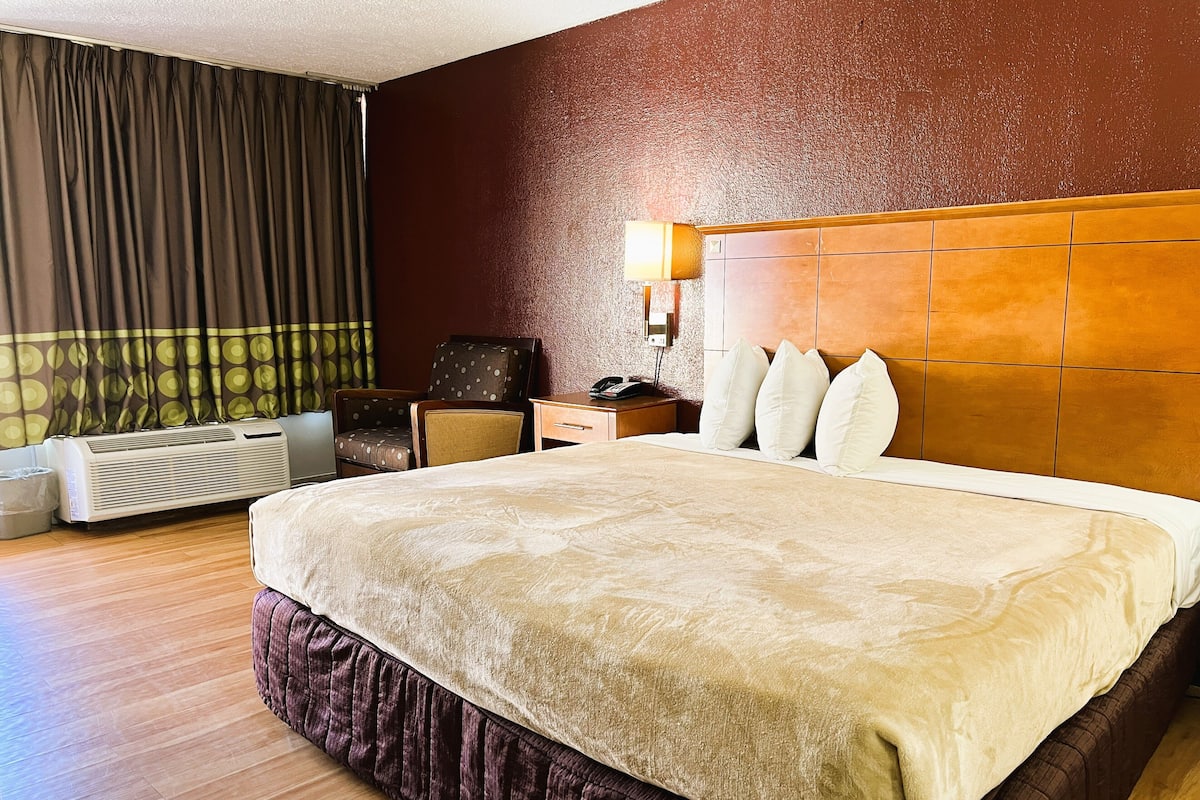 Habitación de hotel con una cama grande, cabecera de madera, mesita de noche con lámpara, una silla y un equipo de aire acondicionado.