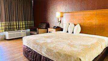 A hotel room with a large bed, a wooden headboard, a nightstand with a lamp, a chair, and an air conditioning unit.