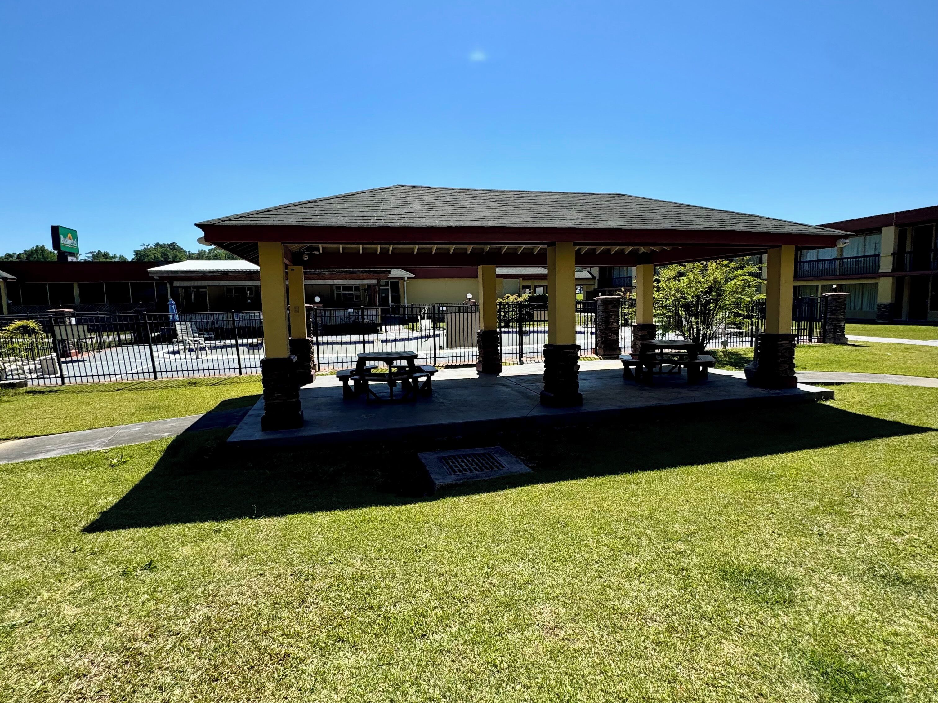 A covered outdoor seating area with picnic tables and benches, surrounded by a fence and a grassy lawn.