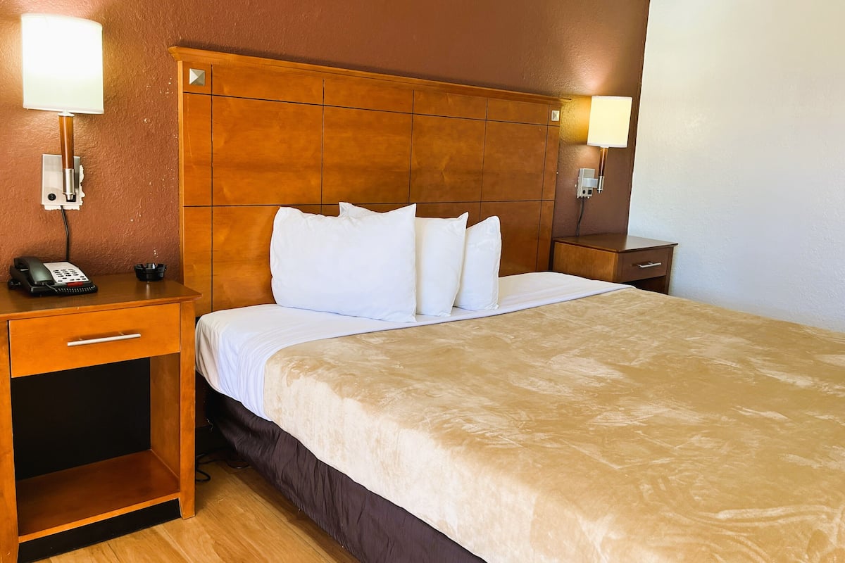 Una habitación de hotel con cabecera de madera, cama con ropa blanca, mesita de noche con lámpara y teléfono en el buró.