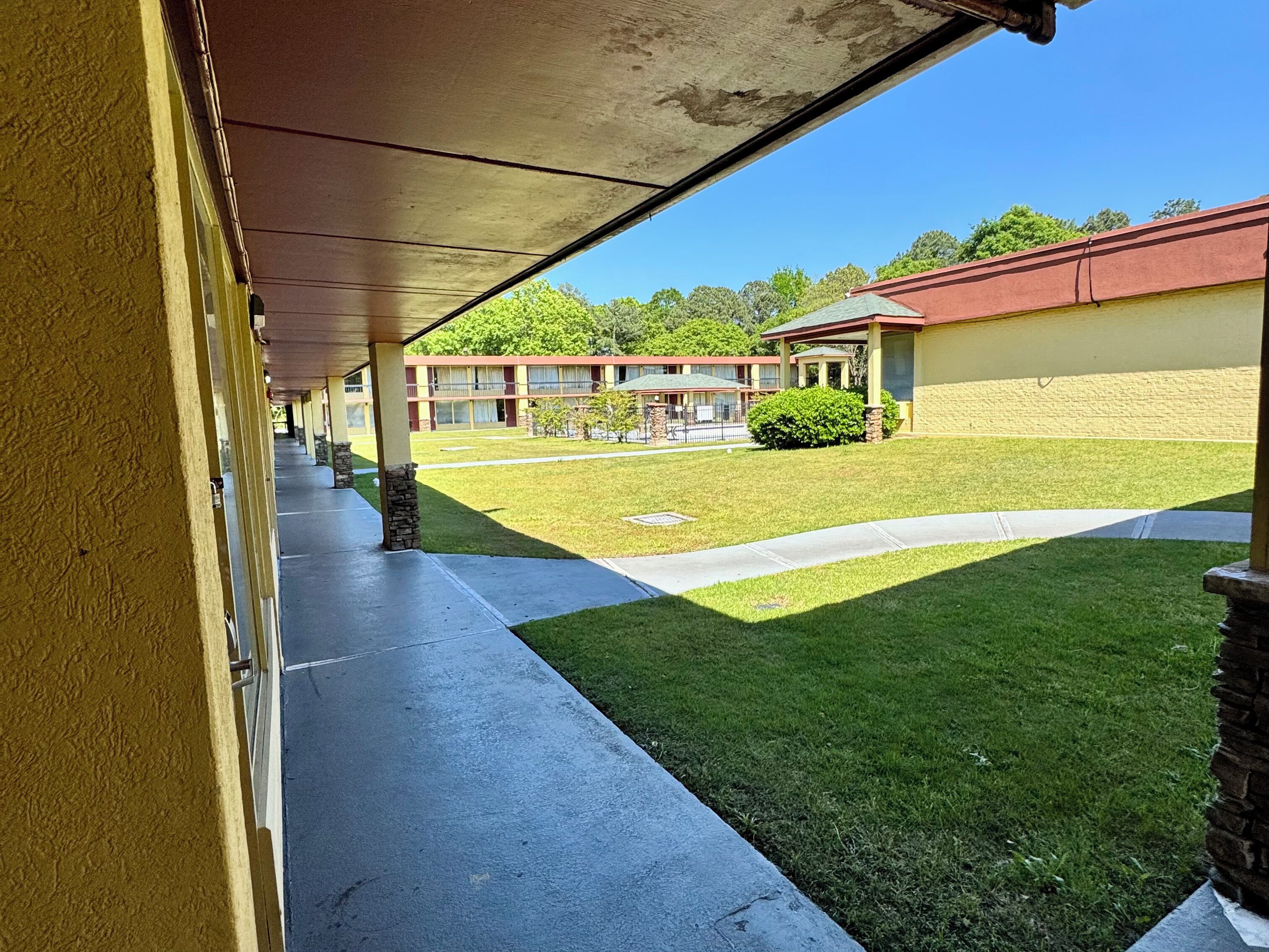 A covered walkway leading to a building with a red roof, a well-maintained lawn, and a clear blue sky.