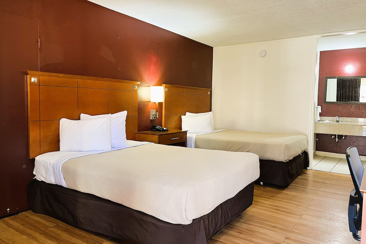 Habitación de hotel con dos camas, cabecero de madera, mesita de noche con lámpara y baño con espejo y lavabo.