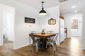 A dining area with a wooden table and four chairs, a pendant light, a wall-mounted mirror, and a refrigerator in the background.