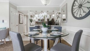 Un coin repas avec une table en verre, des chaises à revêtement gris, un vase blanc orné d’orchidées et une grande horloge murale.