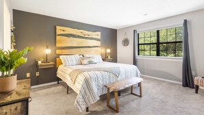 A bedroom with a bed, bedside tables, a bench, a large window, and a wall art piece.