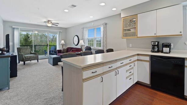 A modern kitchen with white cabinetry, a dishwasher, and a coffee maker. There is a dining area with a table and chairs, and a balcony with a view of trees.