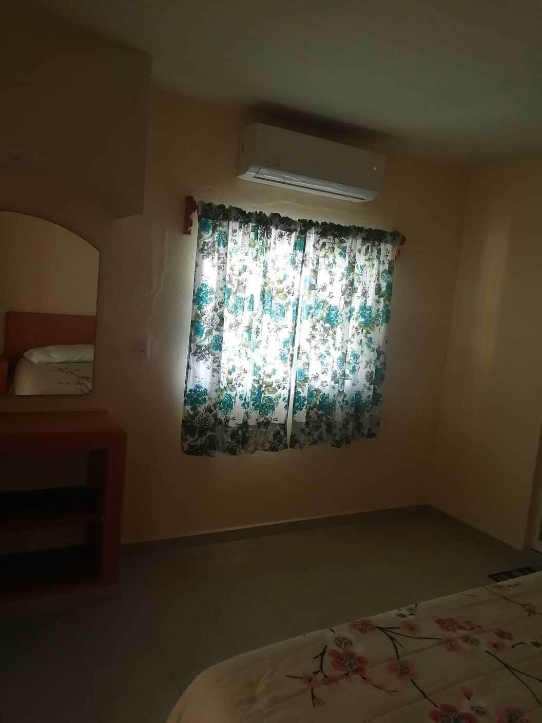 Une chambre à coucher avec un lit, une chaise, une fenêtre ornée de rideaux à motifs floraux et un appareil de climatisation fixé au mur.