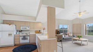 Una cocina moderna con electrodomésticos blancos, una zona de comedor con sofá y mesa de centro, y amplios ventanales con vistas.