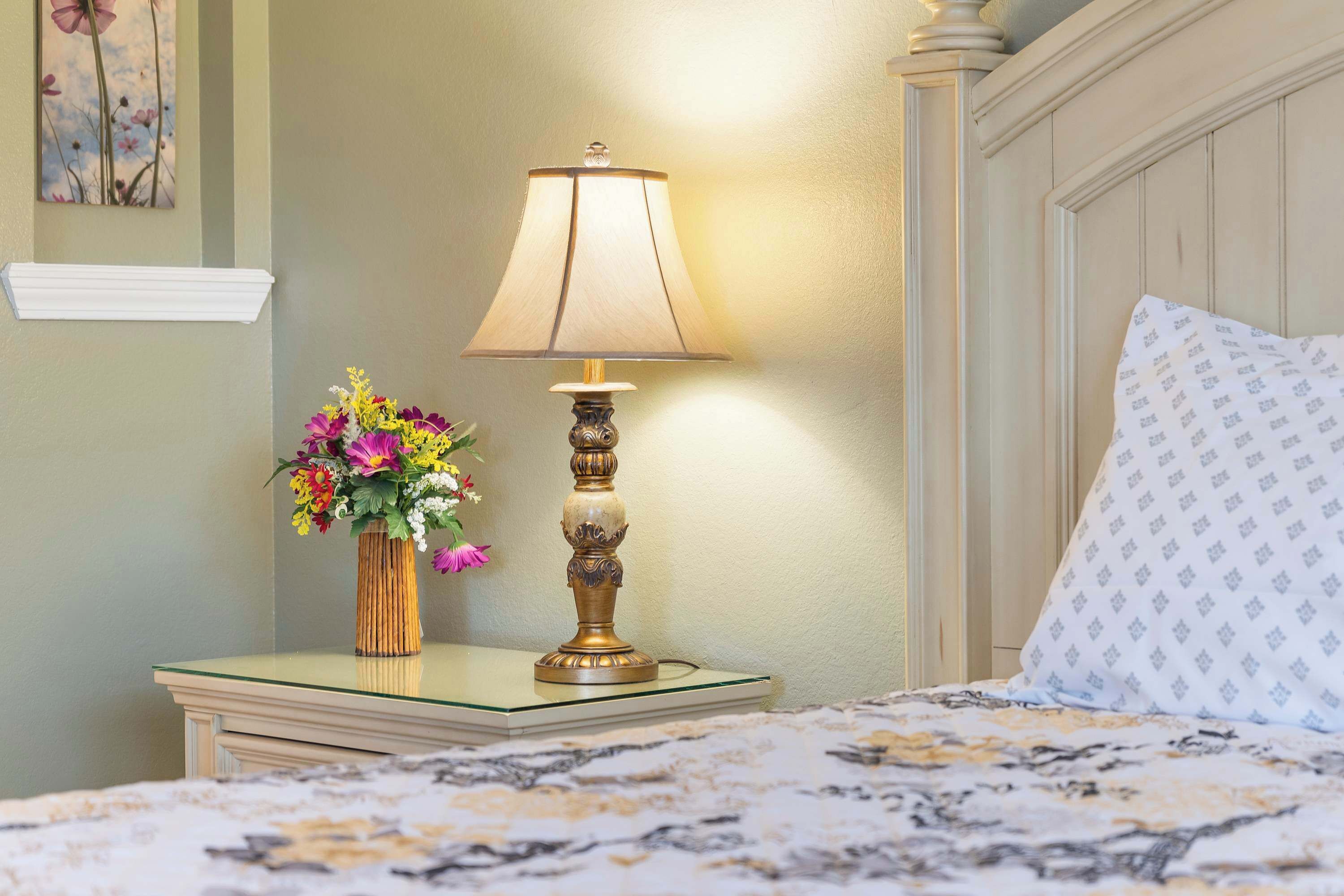 Een net opgemaakt bed met een bedsprei met patroon, een nachtkastje met een lamp en een vaas bloemen, en een muur met een bloemistisch schilderij.