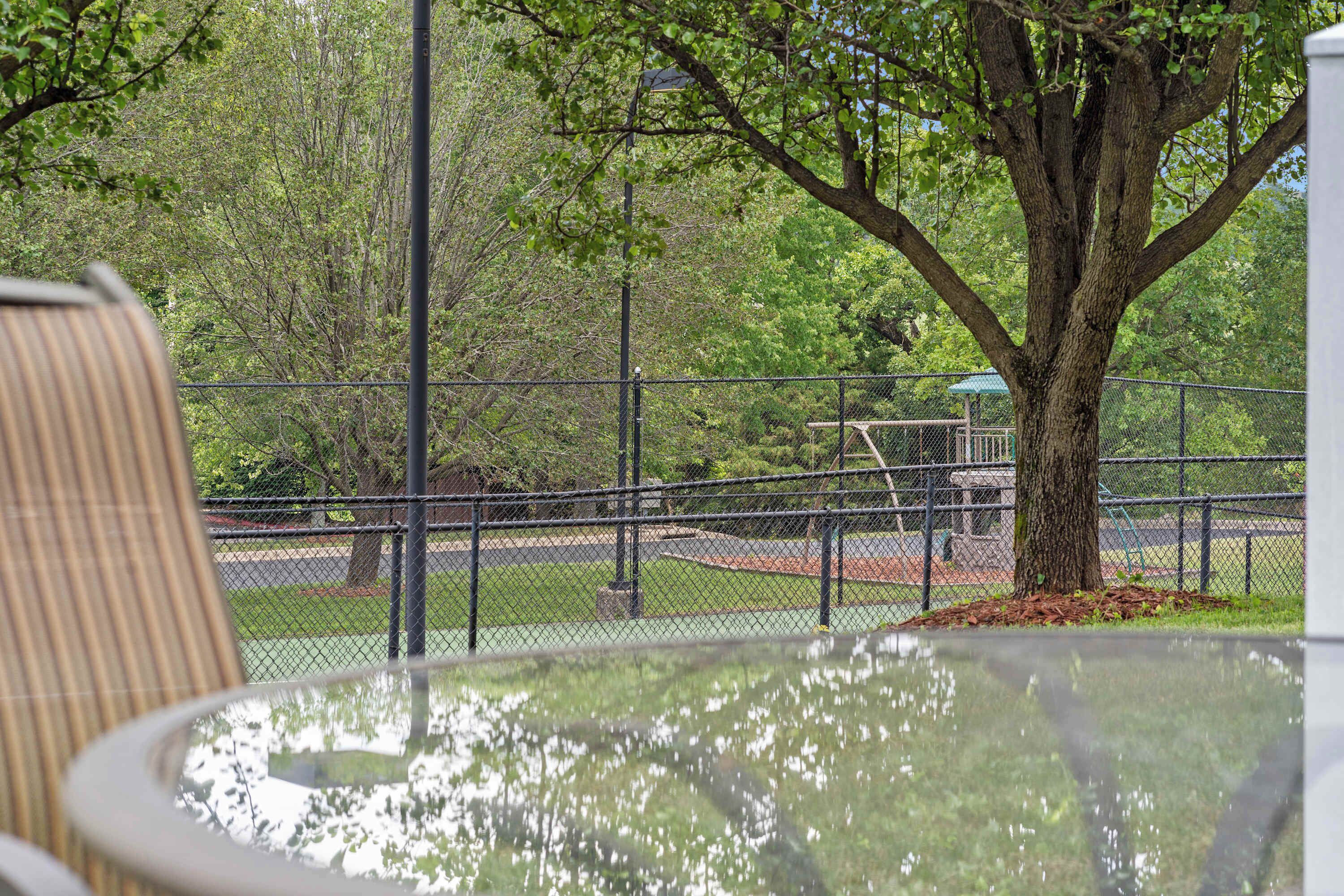A park with a fenced tennis court, a tree, and a bench.