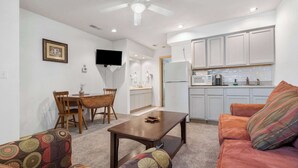 A living room with a sofa, a coffee table, and a TV. There is a kitchen area with cabinets and a refrigerator.