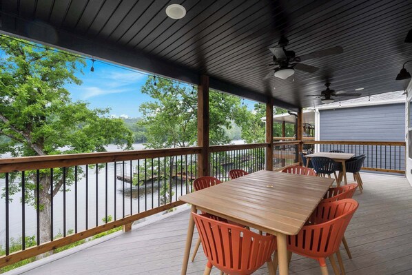 A covered outdoor deck with a wooden table and orange chairs, overlooking a body of water and trees.