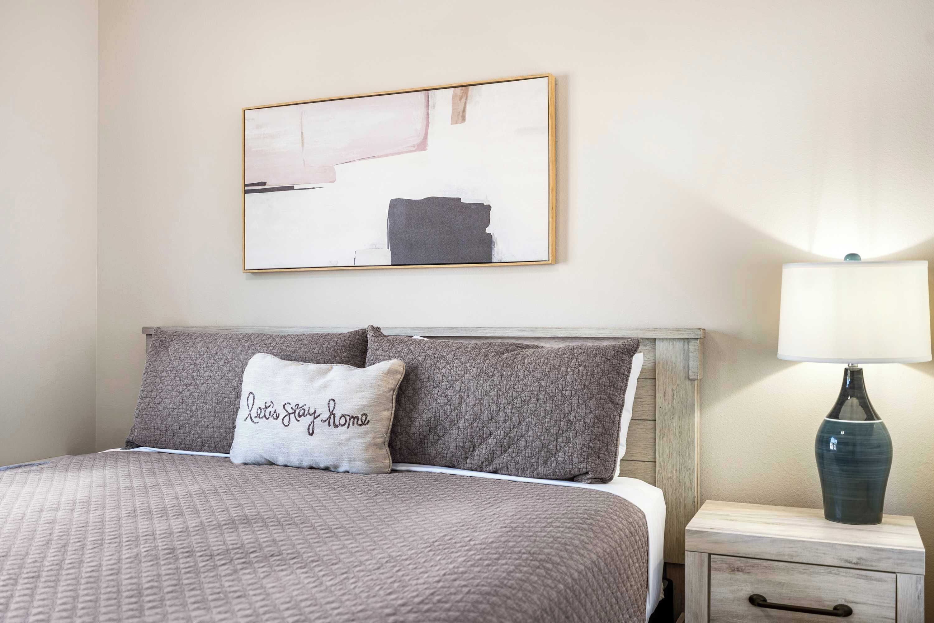 Een net opgemaakt bed met een gewatteerde spreidekbedovertrek, twee kussens, een nachtkastje met een lamp en een lijst met een abstract schilderij aan de muur.