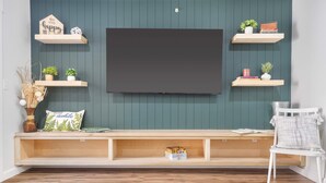 Een woonkamer met een aan de muur bevestigde televisie, houten planken met decoratieve objecten, een bank met boeken en een witte stoel.