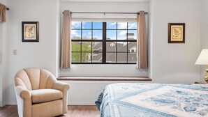 A bedroom with a large window, a beige armchair, a patterned bedspread, and two framed pictures on the walls. - The Greens Escape in Branson | Near DT! (Branson)