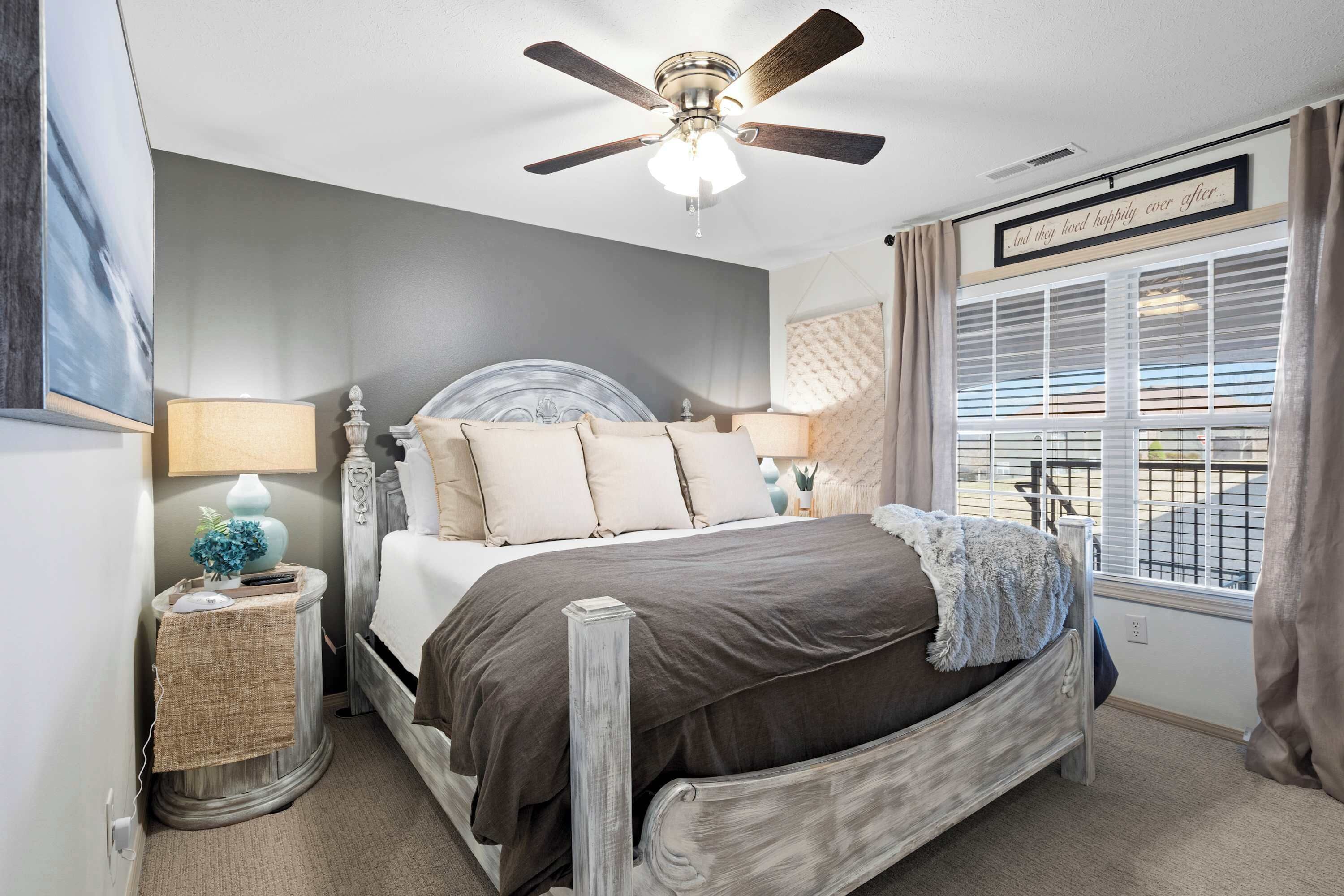 A bedroom with a bed, a ceiling fan, a window with curtains, and a bedside table with a lamp.