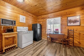 Interior de una cabaña de madera con microondas, estufa, refrigerador y una mesa de comedor con sillas.