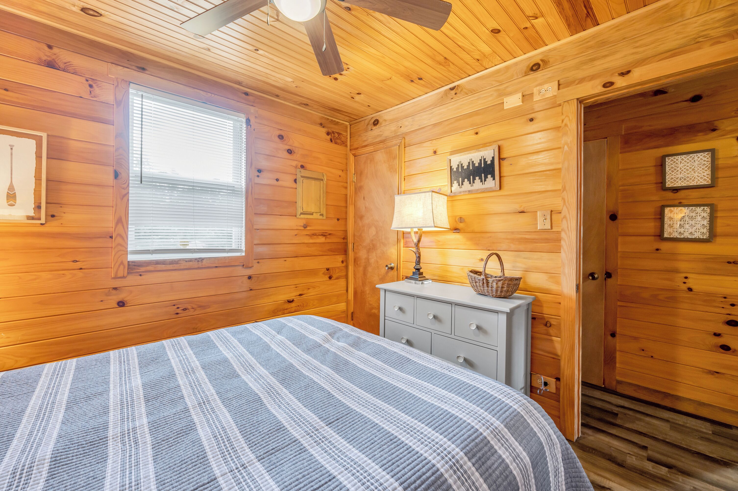 Un dormitorio de cabaña de madera con cama, cómoda, lámpara y ventana.