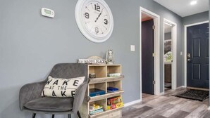 Un pasillo con pared gris, un reloj blanco, una silla gris, un estante de madera con cajas de almacenamiento y una puerta negra.