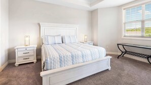 Un dormitorio con una cama grande, mesitas de noche blancas, un banco y una ventana con persianas.