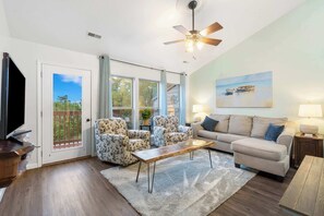 A living room with a sofa, two armchairs, a coffee table, a TV, a ceiling fan, and a painting on the wall.