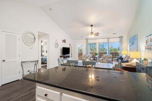 A kitchen with a large island, a dining area with a table and chairs, a ceiling fan, and a view of the outdoors through large windows.