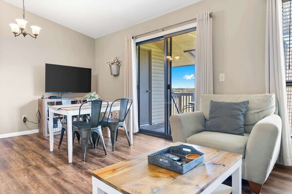 A living room with a TV, dining table, chairs, a sofa, and a sliding glass door leading to a balcony.