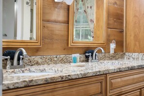 Une salle de bain dotée d’un comptoir en granit, de deux lavabos, d’armoires en bois et d’un miroir agrémenté d’un rideau à motifs floraux.