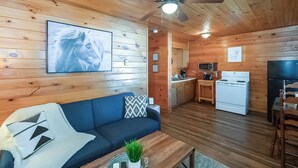 A cozy cabin interior with a blue sofa, wooden walls, a ceiling fan, a kitchen area, and a dining table.