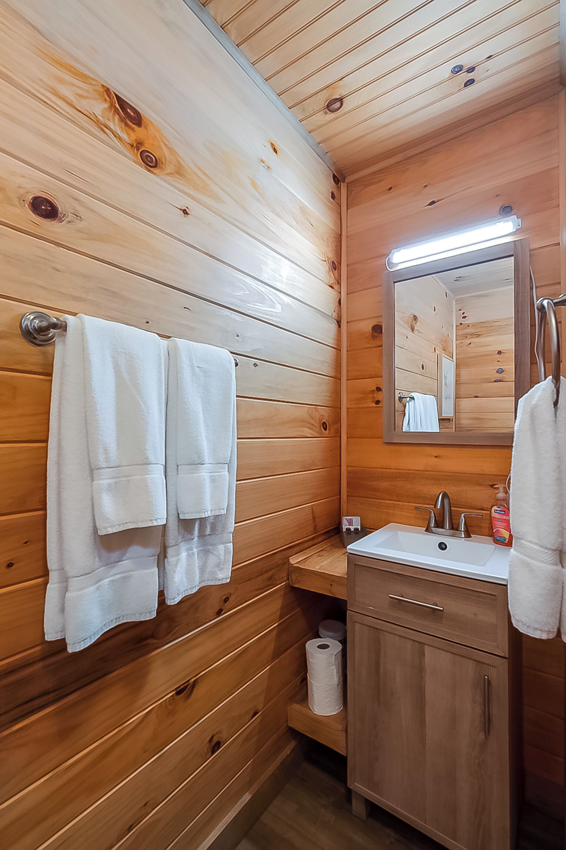 A wooden-paneled bathroom with two towels hanging on a rack, a sink with a mirror, and a toilet with a roll of toilet paper.