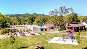 A park with picnic tables, playground equipment, and buildings in the background.
