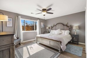 Une chambre à coucher avec un lit, un ventilateur de plafond, des tables de chevet avec des lampes, une fenêtre avec des rideaux et une vue sur les arbres à l’extérieur.