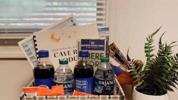 Un panier-cadeau contenant des bouteilles d’eau, des en-cas et une carte, posé sur une table en bois près d’une fenêtre munie de stores.