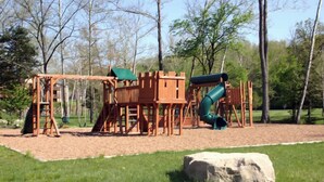 A playground with a wooden play structure, including a slide and swings, set in a grassy area with trees in the background. - Birdie Lane Beauty – Golf, Pools, Gym, & More! (Branson West)