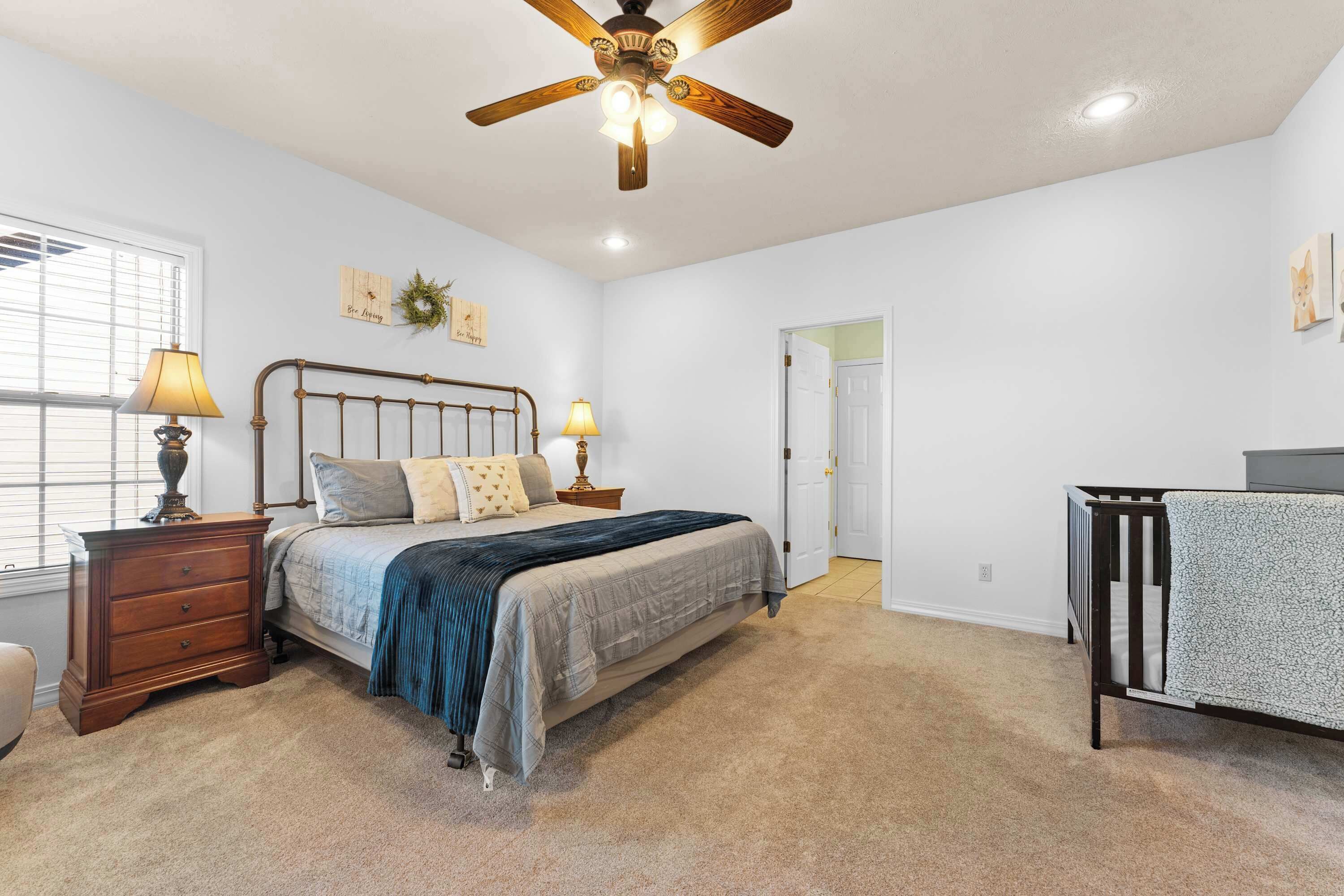 A bedroom with a bed, nightstands, a crib, a ceiling fan, and a door.
