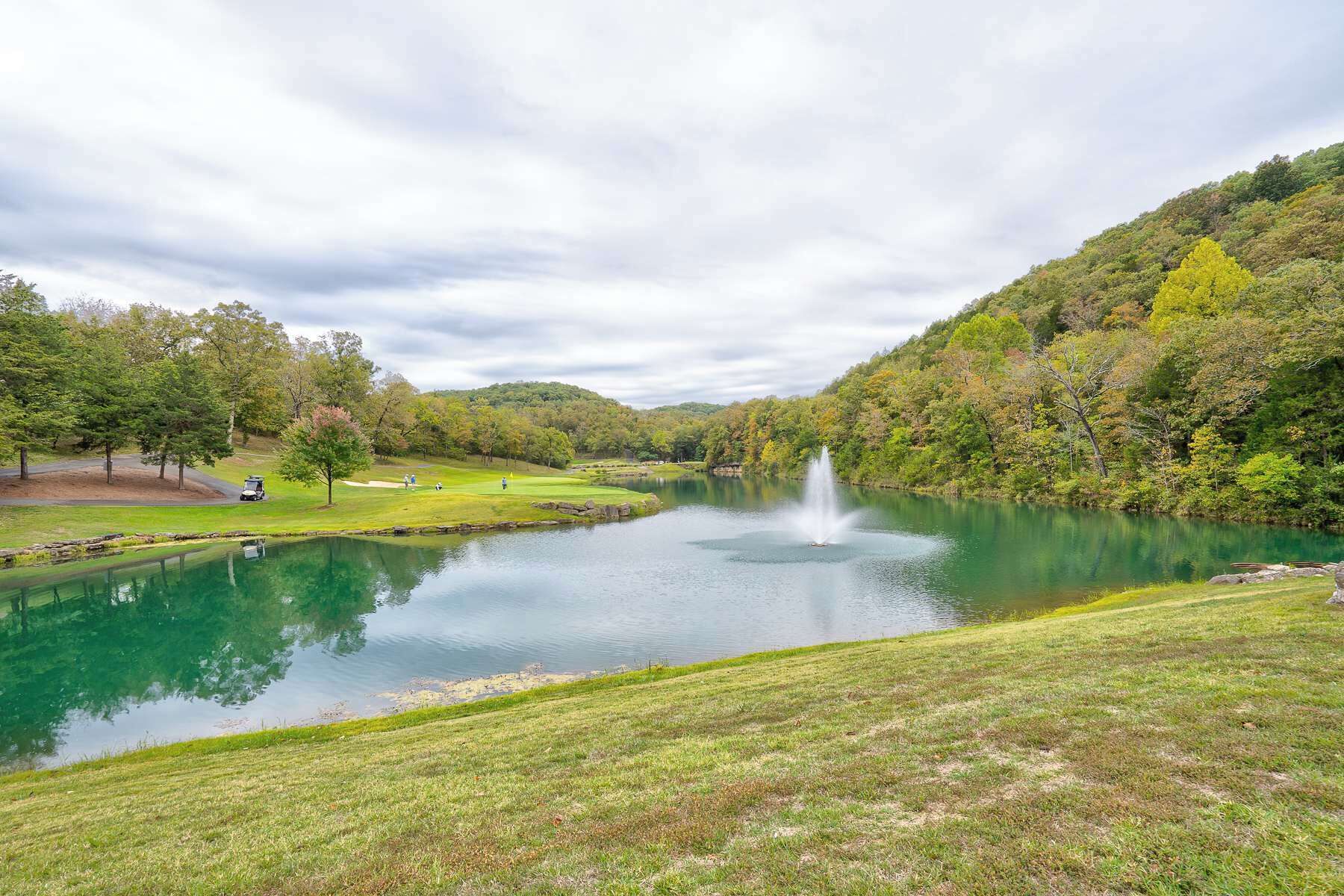 A serene lake with a fountain, surrounded by lush greenery and a golf course.