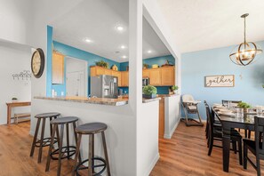A modern kitchen with wooden cabinets, a central island with stools, and a dining area with a chandelier.