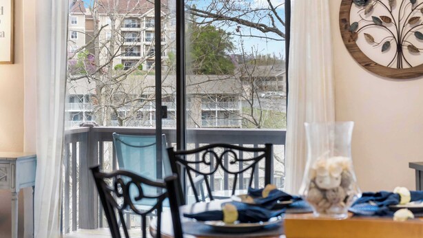 Un comedor con una mesa lista para dos, un florero con adornos y vistas a un área residencial a través de una ventana.