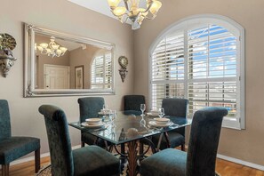 Uma sala de jantar com mesa de vidro, seis cadeiras, lustre, espelho grande e janela com persianas.