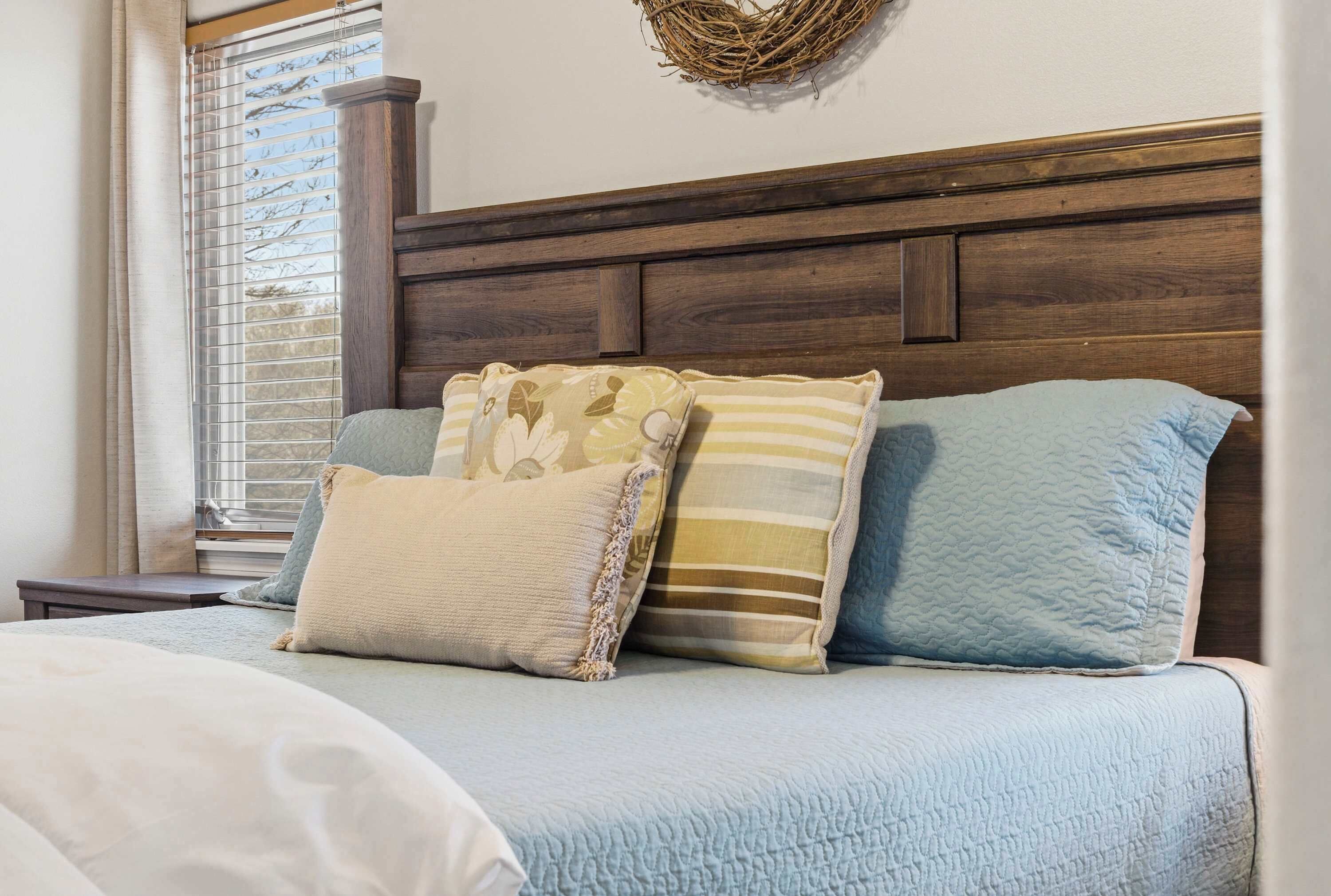A neatly made bed with a blue quilt and multiple pillows, a wooden headboard, and a window with blinds.