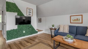 A modern living room with a flat-screen TV, a blue sofa, a wooden coffee table, and a staircase with green foliage.